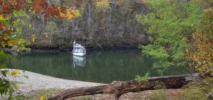 flat calm green lake with a boat at anchor and lines to shore surrounded by trees and fall coloured leaves