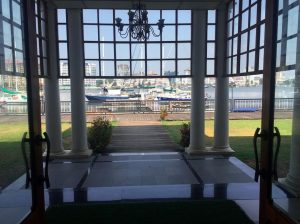 Viewing a marina with boats docked through an entrance way with high chandelier and windows, pillars and polished floor