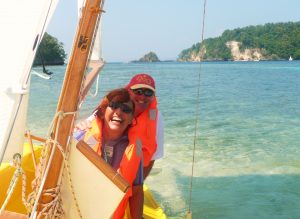 a smiling lady in a white t shirt with a red cap sitting on a large white sail on a pontoon making repairs