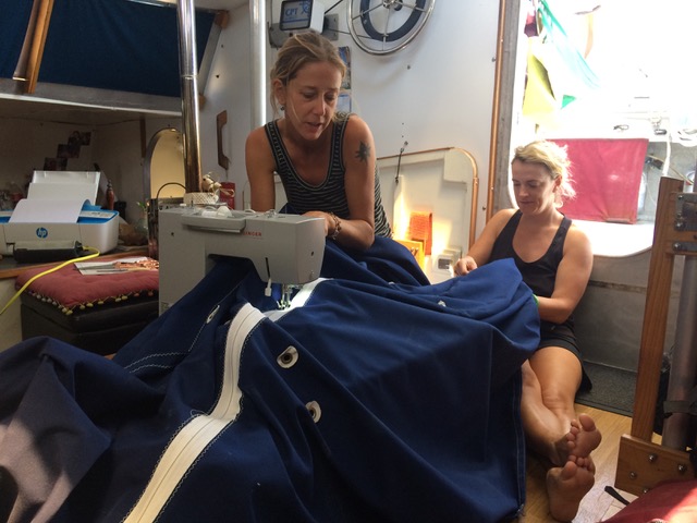 inside the boat cabin a woman sits up against the hatchway holding a large piece of sail material and in the foreground another woman works the material through a big sewing machine